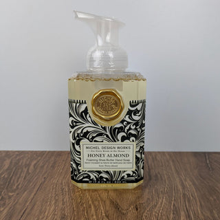A pump bottle of Honey Almond Foaming Soap by Michael Design Works, with a black and white floral pattern on the label and a gold wax seal.
